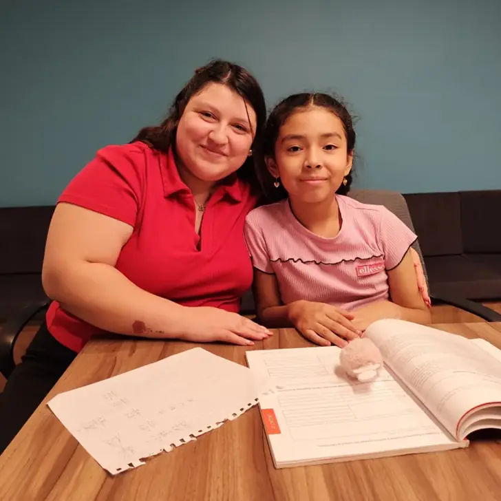 Profesora mirando con orgullo a su alumna mientras ambas sonríen frente a su material de trabajo.
