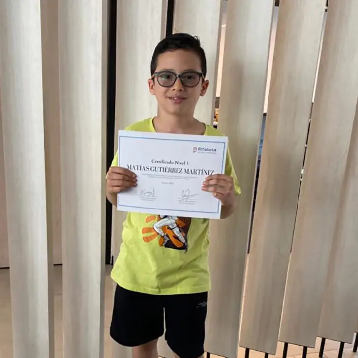 Niño pequeño sosteniendo con orgullo su diploma de Alfabeta, sonriendo ante la cámara en un ambiente escolar festivo.